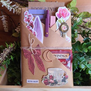 May include: A brown paper package tied with twine, decorated with stickers, a teacup, and a tea bag. The package includes a bookmark, a pen, and a crescent moon charm. The text "BOOK REVIEW" is visible.