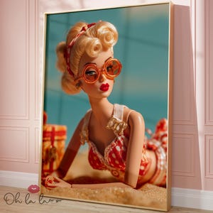 以下が含まれることがあります： レトロスタイルの人形が描かれたフレームプリント。人形はブロンドの髪、赤い口紅、オレンジ色のサングラスをしています。赤と白のドット柄の水着を着ています。背景は水色で、金色のフレームが付いています。