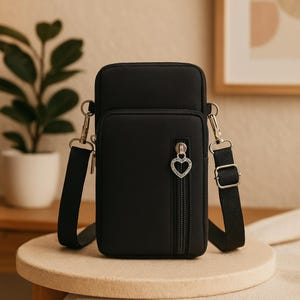 May include: Black crossbody bag with a front zippered pocket and a decorative heart-shaped zipper pull. The bag has a black adjustable shoulder strap and silver-colored hardware. The bag is sitting on a light-colored surface.