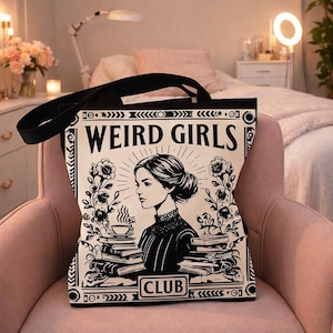 Peut inclure: Sac fourre-tout noir avec le texte "WEIRD GIRLS CLUB" dans un style vintage. Le design présente un portrait de femme, des livres, des fleurs et une tasse de café, le tout en noir et blanc. Le sac a des poignées noires et est posé sur une chaise rose.