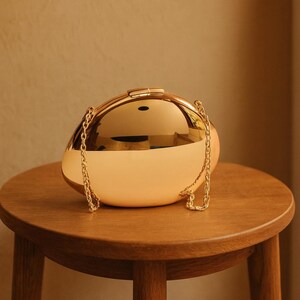 May include: A gold-coloured, egg-shaped clutch bag with a matching chain strap. The bag has a shiny, reflective surface and a clasp closure. It sits on a small, round wooden table.