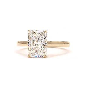 Può includere: Un anello d'oro con un grande diamante a taglio rettangolare. Il diamante è il punto focale, incastonato in una fascia semplice ed elegante. L'anello è presentato su uno sfondo bianco, evidenziando la sua brillantezza e il suo design.