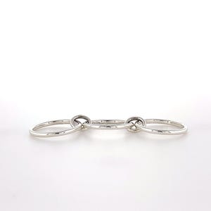 May include: A set of three interlocking silver rings arranged in a horizontal row. The rings are smooth, polished, and circular, connected by two smaller silver jump rings that link the central ring to the two outer rings, creating a continuous chain.