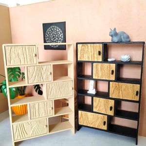 Può includere: Due librerie in legno in miniatura. Una è beige chiaro con ante e ripiani con texture effetto legno. L'altra è nera con ante con texture effetto legno. Una statuina di gatto grigio è appoggiata sopra la libreria nera.