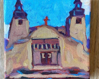 May include: An oil painting on canvas depicts a church with two bell towers and a red cross on the roof. The artwork uses a palette of blues, yellows, and purples to create a textured, impressionistic style. The canvas is square and mounted on a wooden surface.