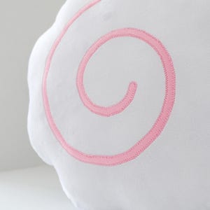May include: A white, cloud-shaped plush pillow with a pink, embroidered spiral design. The soft, fluffy pillow is a decorative item, likely for a child's room or nursery. The pink stitching provides a contrasting accent to the white fabric.