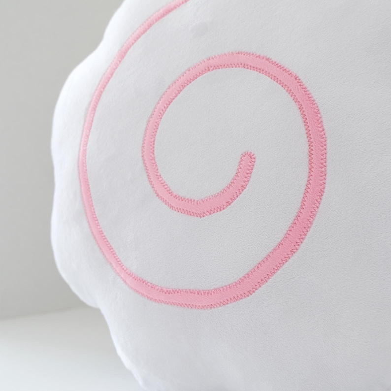 May include: A white, cloud-shaped plush pillow with a pink, embroidered spiral design. The soft, fluffy pillow is a decorative item, likely for a child's room or nursery. The pink stitching provides a contrasting accent to the white fabric.