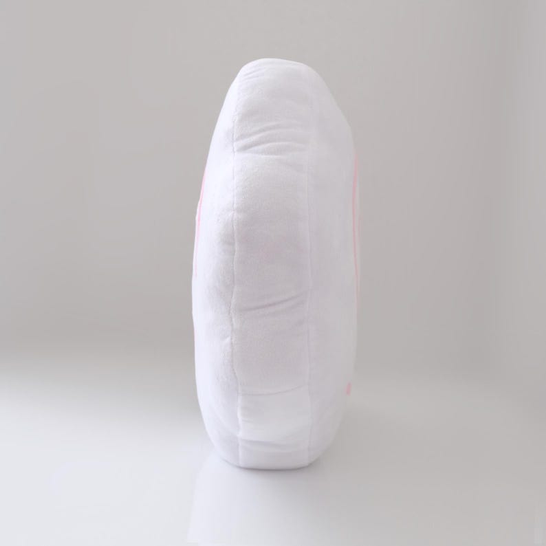 May include: A white plush pillow with a rounded shape. The pillow has a soft, textured surface and a pink design on the side. The pillow is designed for comfort and support.