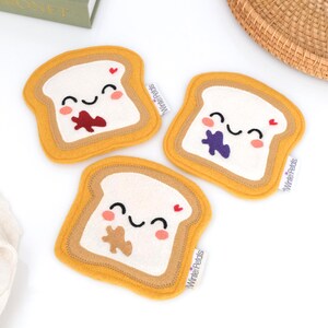 May include: Three felt coasters shaped like slices of bread with smiling faces and different fillings: peanut butter, jelly, and chocolate. Each coaster has a yellow crust, a white center, and a small red heart. The text "Winnie Petals" is printed on each coaster.