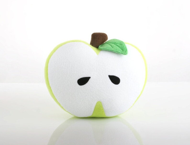 May include: A green plush apple pillow with a white face and black eyes. The apple has a brown stem and a green leaf.