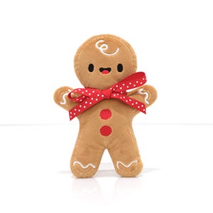 May include: A gingerbread man plush toy with a red and white polka dot bow tie. The gingerbread man is smiling and has two red buttons on its chest.