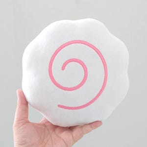 Könnte beinhalten: Ein weißes, rundes Plüschkissen in Form eines Narutomaki-Fischkuchens mit einem markanten, aufgestickten rosa Spiralmuster in der Mitte. Das weiche, abgerundete Objekt hat einen gewellten Rand und eine flauschige Textur auf der gesamten Oberfläche.