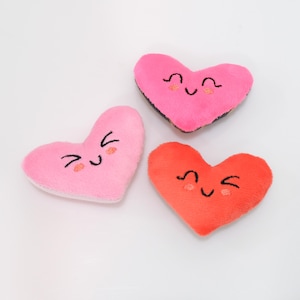 Peut inclure: Trois jouets en peluche en forme de cœur avec des visages souriants. Les cœurs sont roses, rouges et rose clair.