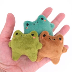May include: Three small, plush frog toys in shades of green and brown. Each frog has black eyes, a smiling mouth, and pink cheeks. The frogs are soft and cuddly, suitable for children or as decorative items.