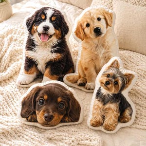 May include: Four plush dog-shaped pillows in various breeds: a Bernese Mountain Dog, a Golden Retriever, a Dachshund, and a Yorkshire Terrier. The pillows are on a cream-colored bedspread, showcasing the soft, detailed designs of the puppies.
