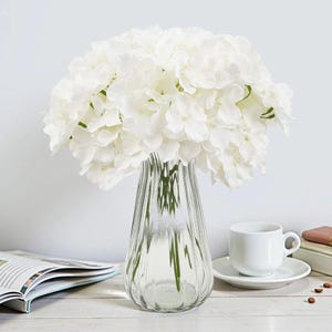 Op de afbeelding: Een heldere glazen vaas met verticale ribbels bevat een groot boeket witte hortensia's. Rechts staan een witte kop en schotel, met een boek en een kleine stapel boeken in de buurt. De opstelling staat op een wit oppervlak.