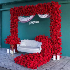 May include: A decorative floral arch and sofa arrangement. The arch is covered in red roses, framing a white and pink fabric drape and the word "JAROWN" in neon. A white sofa with a single pillow is centered below the arch, surrounded by more red roses.