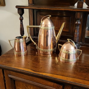 May include: A copper tea set with brass accents, including a teapot, creamer, and sugar bowl. The set is displayed on a dark wooden surface, showcasing its polished finish and classic design. The teapot has a tall, slender shape.