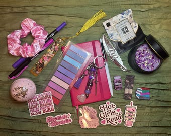 May include: A collection of pink and purple items, including a scrunchie, pens, stickers that say "Hot Girls Read" and "Bookish Baddie", a notebook, a bath bomb, and a candle. There is also a package of white tea and a decorative feather.