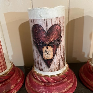 May include: A decorative candle holder with a distressed red base and a cylindrical body. The body features a heart design with the words "Love Never Fails" on a tag. The background has a rustic, weathered wood appearance.