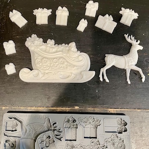 May include: White resin Christmas decorations including a sleigh, reindeer, and several gift boxes. A gray mold with the text "SANTA'S SLEIGH IOD" is also present, featuring similar designs for crafting.