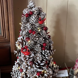 May include: A decorative Christmas tree made of pine cones, painted white with red berries and small red bird ornaments. The tree is illuminated with small lights and sits on a wooden base. A festive holiday decoration.