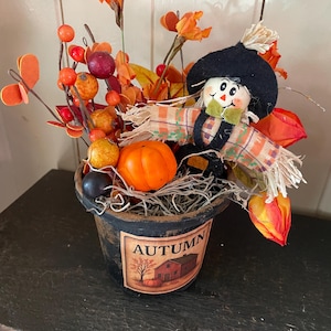 May include: A decorative autumn-themed arrangement in a black pot. It features a small scarecrow figure, orange berries, flowers, and a miniature pumpkin. The pot has a label with the word "AUTUMN" and a picture of a house and tree.