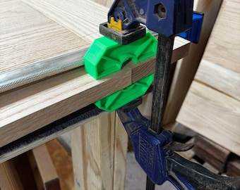 May include: A blue Irwin bar clamp securing two pieces of light-colored wood together. The clamp features bright green plastic corner protectors positioned at the joint where the wood pieces meet, providing pressure to hold the boards in place.