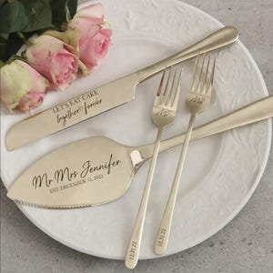 Puede incluir: Un juego de servir pastel de bodas plateado con cuchillo, pala y dos tenedores. El cuchillo dice "LET'S EAT CAKE together forever". La pala está grabada con "Mr Mrs Jennifer EST DECEMBER 31, 2022". Los tenedores están marcados "Mr" y "Mrs" con la fecha "12.31.22".