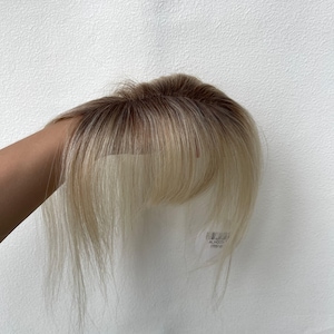 May include: A hair topper featuring a rooted design with dark brown roots transitioning into light blonde, platinum-toned strands. The piece includes a sheer lace base at the crown and a set of wispy, face-framing bangs that fall straight.