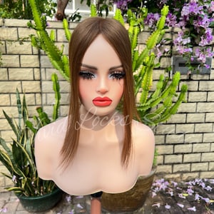 May include: A mannequin head with a brown wig. The mannequin has full makeup, including dark eyelashes and red lipstick. The text "Wild Locks Sun" is visible on the mannequin. The background includes plants and a brick wall.