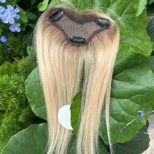 May include: A blonde hair topper with a brown mesh base and several attached clips. The hair is a light blonde color with darker roots. The topper is laying on a bed of green leaves and flowers.