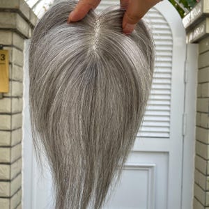 Puede incluir: Una pieza de cabello gris con una raya de aspecto natural. La pieza de cabello se sostiene, mostrando los finos mechones y la forma en que se asentaría en la cabeza. El color es una mezcla de tonos grises, dando una apariencia realista.