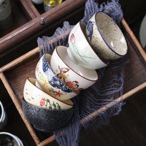 Pode incluir: Uma pilha de tigelas de cerâmica com vários desenhos florais e de pássaros. As tigelas são brancas, bege e cinza escuro, com detalhes pintados à mão em vermelho, azul e amarelo. Estão dispostas numa bandeja de madeira com um pano azul.