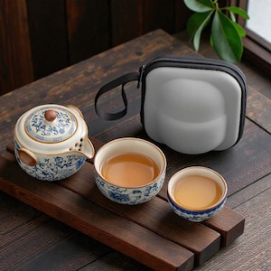 May include: A traditional tea set featuring a teapot, two teacups, and a grey carrying case. The teapot and cups display a blue and white floral pattern. The teacups are filled with tea. The set is on a wooden tray.