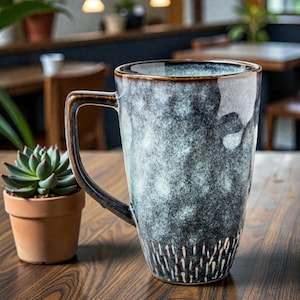 May include: A tall, speckled ceramic mug with a dark blue and grey glaze. The mug has a large handle and a brown rim. The bottom of the mug features a white, vertical line design. A small succulent plant in a terracotta pot sits to the left of the mug.