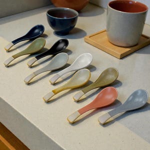 Puede incluir: Una colección de cucharas de sopa de cerámica en varios colores, incluyendo azul marino, verde oliva, negro y melocotón. Cada cuchara tiene un mango blanco y un acabado moteado. Un cuenco azul y una taza están en el fondo.
