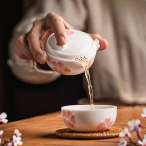 May include: A white ceramic teapot and teacup set adorned with pink floral patterns. Tea is being poured from the teapot into the teacup. The teapot features a lid with a flower-shaped handle. The teacup rests on a small, round coaster.
