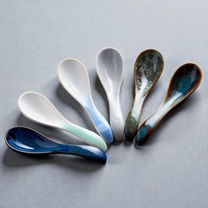 May include: A collection of six ceramic spoons with a glossy finish. Each spoon features a unique color gradient, including white, blue, green, and gray. The spoons are arranged on a gray surface, showcasing their varied designs and textures. Ideal for serving small portions.