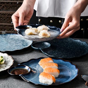 May include: A collection of blue and gray ceramic sushi plates with a unique wavy design. Some plates hold sushi and dipping sauce. The plates are speckled with a textured finish, and are being held by a person.