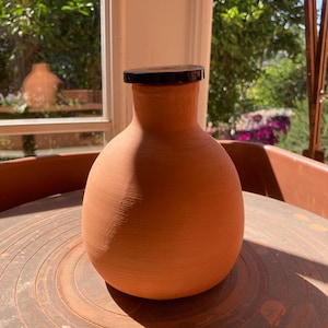 Peut inclure: Un vase en terre cuite fait à la main avec un couvercle marron foncé. Le vase a un corps arrondi et un col étroit. La terre cuite est d'une couleur orange chaude et terreuse. Le vase est sur un tour de potier.