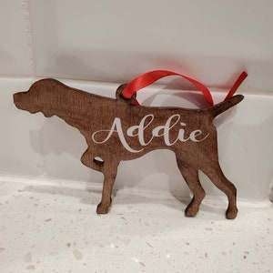 May include: A wooden dog-shaped ornament with the name "Addie" in white script. The ornament is a medium brown color and has a red ribbon for hanging. The dog is in a standing pose.