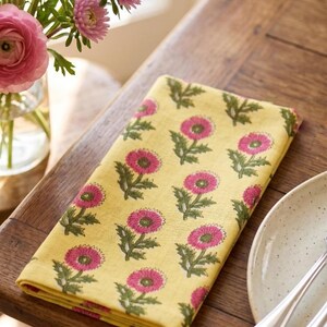 Puede incluir: Una servilleta cuadrada doblada de color amarillo pálido con un patrón repetitivo de flores rosas con tallos y hojas verdes. La tela muestra un estampado floral consistente en toda la superficie, exhibiendo un diseño botánico vibrante.