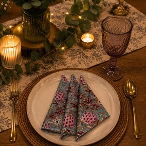 May include: Three folded fabric napkins with a teal background and a repeating floral pattern in pink, white, and brown, placed on a cream-colored ceramic plate. The plate sits on a round woven charger, flanked by a gold fork and a gold spoon.