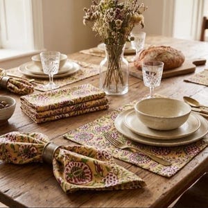 May include: A dining table setting featuring rectangular placemats and matching cloth napkins in a yellow fabric with a pink and green floral pattern. The napkins are secured with metallic rings, accompanied by ceramic bowls, plates, and gold cutlery.