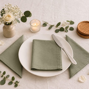 Pode incluir: Um prato de cerâmica branca com um guardanapo de tecido verde sálvia dobrado e uma faca de metal prateado. Dois guardanapos verde sálvia adicionais estão colocados nas laterais, acompanhados por um pequeno vaso com uma rosa branca.