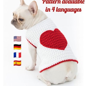 May include: A white crochet dog sweater featuring a red heart. The sweater, trimmed in red, is worn by a white French Bulldog. The image includes text stating "Pattern available in 4 languages".
