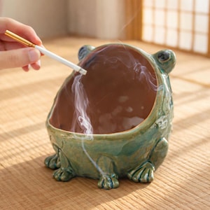 May include: A ceramic ashtray shaped like a frog, with a green exterior and a brown interior. Smoke rises from the ashtray, and a lit cigarette is held above it. The ashtray has four feet and two small ears.