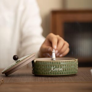 Puede incluir: Cenicero de cerámica verde con un patrón de panal y el nombre "Kevin" en escritura blanca. El cenicero tiene una tapa a juego con un pomo marrón oscuro. Un cigarrillo encendido descansa en el cenicero.