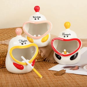 Może przedstawiać: Trzy ceramiczne popielniczki w fantazyjnym kształcie kaczki. Każda popielniczka jest biała z kolorowymi akcentami, w tym czerwonym, żółtym i czarnym. Popielniczki mają otwór w kształcie serca i mały, okrągły guzik na górze. Każda popielniczka ma imię.